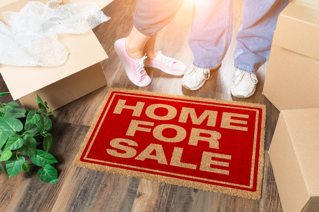 Man and Woman Standing Near Home For Sale Welcome Mat, Moving Boxes and Plant.